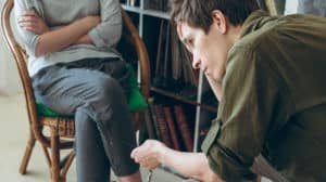 Regretful young man sitting across from his wife who is upset with her arms folded across her chest