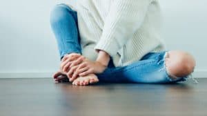 Lower half of a young woman sitting on a floor with a white-walled background. Her hands are wrapped around one of her ankles