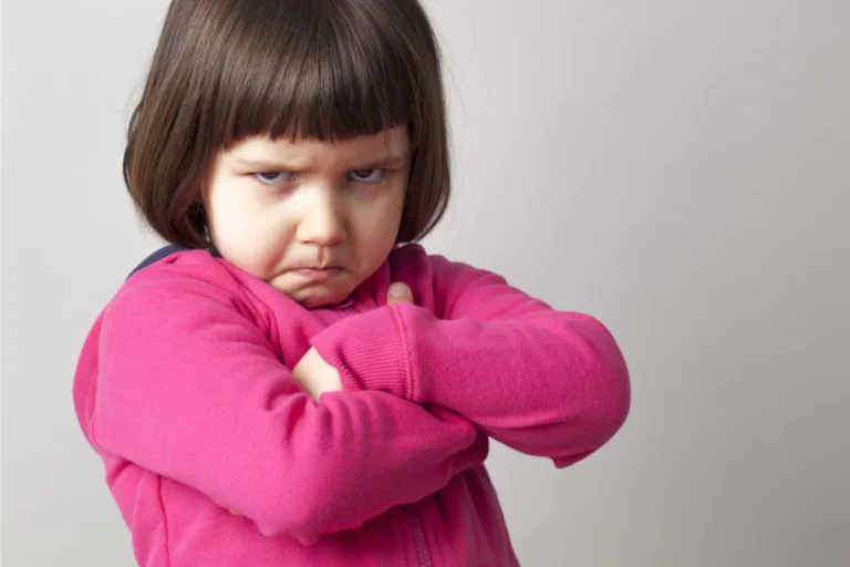 A young girl with a serious expression on her face, arms crossed in defiance, and wearing a bright pink sweater. This image represents a common challenge parents face when dealing with stubborn or defiant behavior and raises questions on how to spank effectively and responsibly.