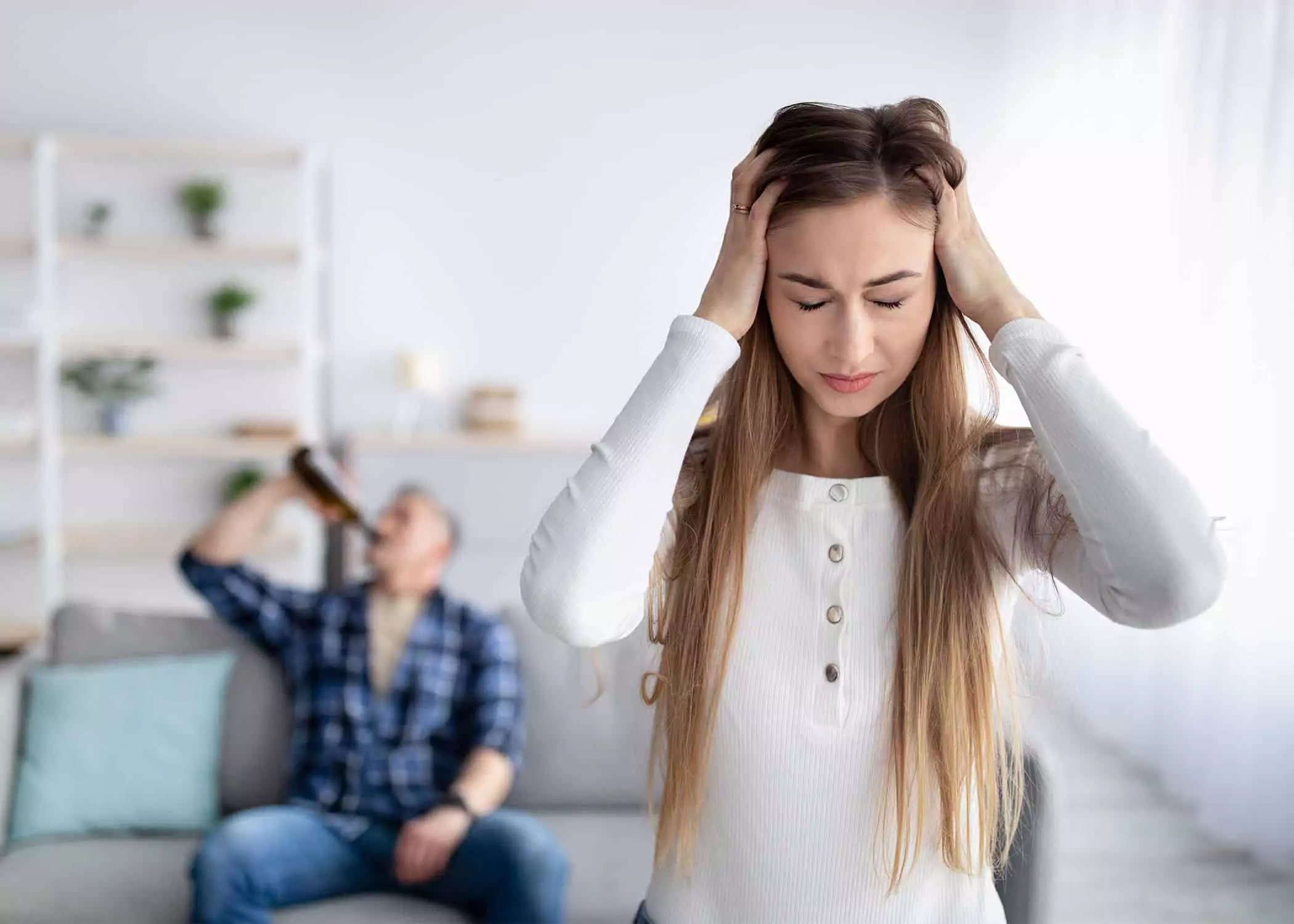 Depressed-wife-married-to-an-addict-husband-drinking-alcohol-from-bottle-in-background