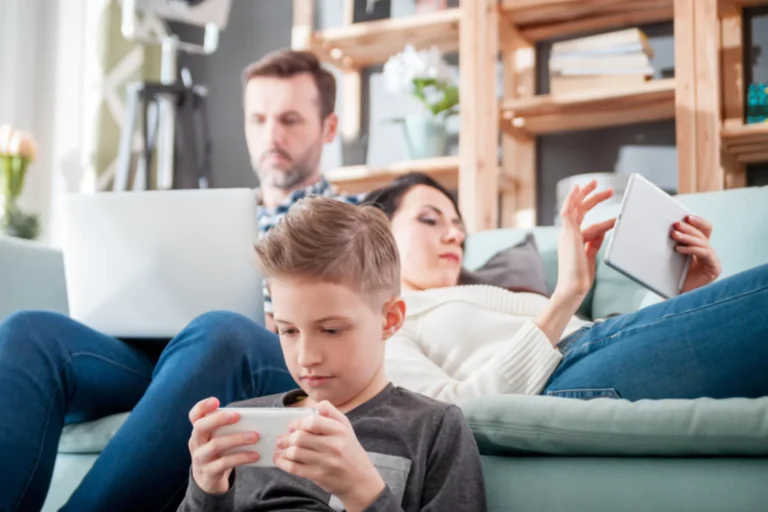 A family sitting on a couch, each absorbed in their own electronic devices: a young boy playing on a smartphone, a woman using a tablet, and a man working on a laptop in the background