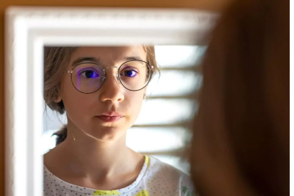 Teenage girl with glasses thoughtfully looking in the mirror